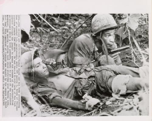 Aid and Protection: pistol drawn and his body protecting that of wounded U.S. paratrooper, medic PFC Andrew G. Brown of Chicago searches jungle for sniper. He and Spec. 4 Gerald Levy of New York crawled through the jungle to dress the wounds of the unidentified paratrooper while a live grenade, ready to go ff at the slightest movement, lay nearby. Action took place yesterday near Thuong Lang, Vietnam