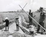 United States marines watched while Vietnamese farmers, using the traditional hand operated equipment, worked in an irrigation ditch of a rice paddy in the Hieu Duc district. The farmers continue to pursue chores despite increasing pressure from the Viet Cong
