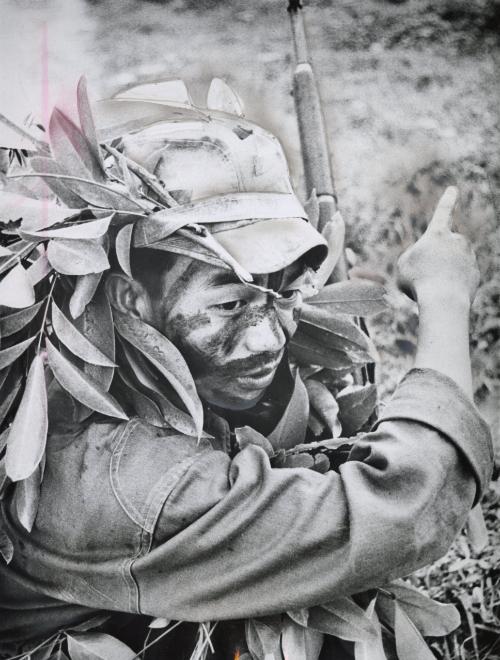 A Royal Laotian soldier, with painted face and leaf trimmed uniform, pointed to an object of suspicion while on a training patrol in the jungle near Vientiane. He was with troops being trained in jungle warfare by American instructors