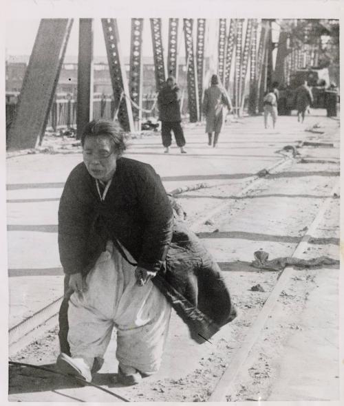 War-Weary Refugee: bent and worn by the shifting tide of war that twice swept over Pyongyang, and elderly North Korean woman plods wearily over the undestroyed span of a Taedong River bridge, as she flees south from the North Korean capital December 4 ahead of the advancing Chinese Reds.