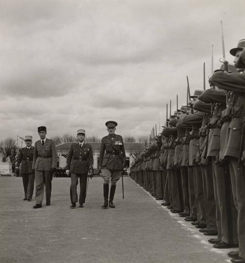 War Turns French Cadets' Fete into Grim Service: For many years the famous French military academy of St. Cyr, the West point of France, has held a gay and colorful yearly fete, known as "Le Triompne de St. Cyr". On this occasion the class of freshmen cadets is solemnly "baptized," receiving the name by which their class will be known. This year there was no rejoicing, no colorful pageant, but a grim parade in wartime uniforms. The cadet class received their name, "Amitie Franco-Britannique ("Franco-British Friendship"). / The cadets are reviewed by the British Military Attache (right), the commander of the academy (center), and the young sub-lieutenant (left), an old St. Cyrian, who performed the batpizing ceremony.