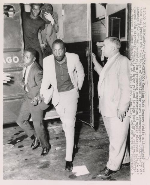 Comedian Dick Gregory (white suit-center) leaves wagon that brought him from House of Correction for court appearance here. Gregory was arrested August 12 during demonstration protesting construction of mobile classroom site. He was charged with disorderly conduct, and later refused to sign bond for his release. Handcuffed to Gregory is James Sanders, man at right is not identified
