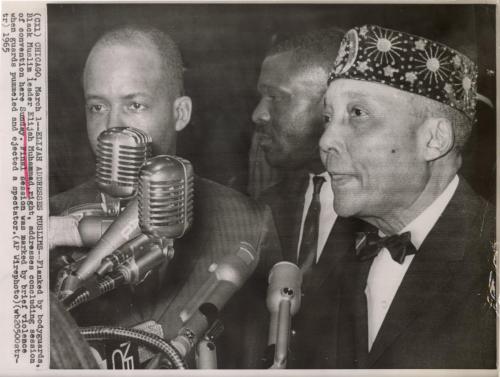 Elijah Addresses Muslims: flanked by bodyguards, Black Muslim leader Elijah Muhammad, right, addresses concluding session of convention. Final session was marked by brief violence when guards pummeled and ejected a spectator, Chicago.