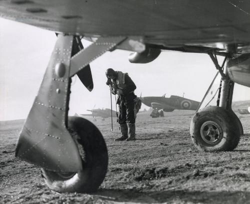 A Hurricane pilot of No. 87 Squadron dons his flying kit, seen from beneath the wings of another aircraft, Exeter