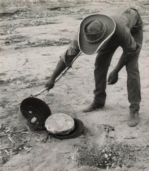 Aboriginal man baking damper (bush bread) in a campoven (cast-iron pot with lid) outdoors by an open fire, Australia, 1968